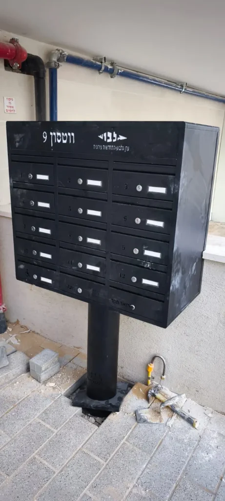 תיבת דואר מעוצבת לבית פרטי עם מספרים, מותקנת על עמוד, בסביבה בנויה.